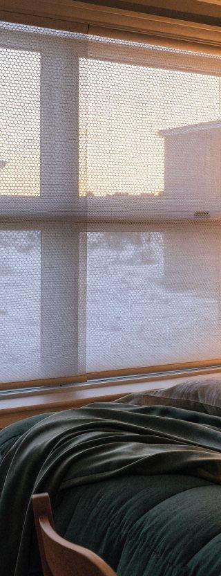 White cellular honeycomb shades half lowered over a bedroom window with snow outside and warm lamplight