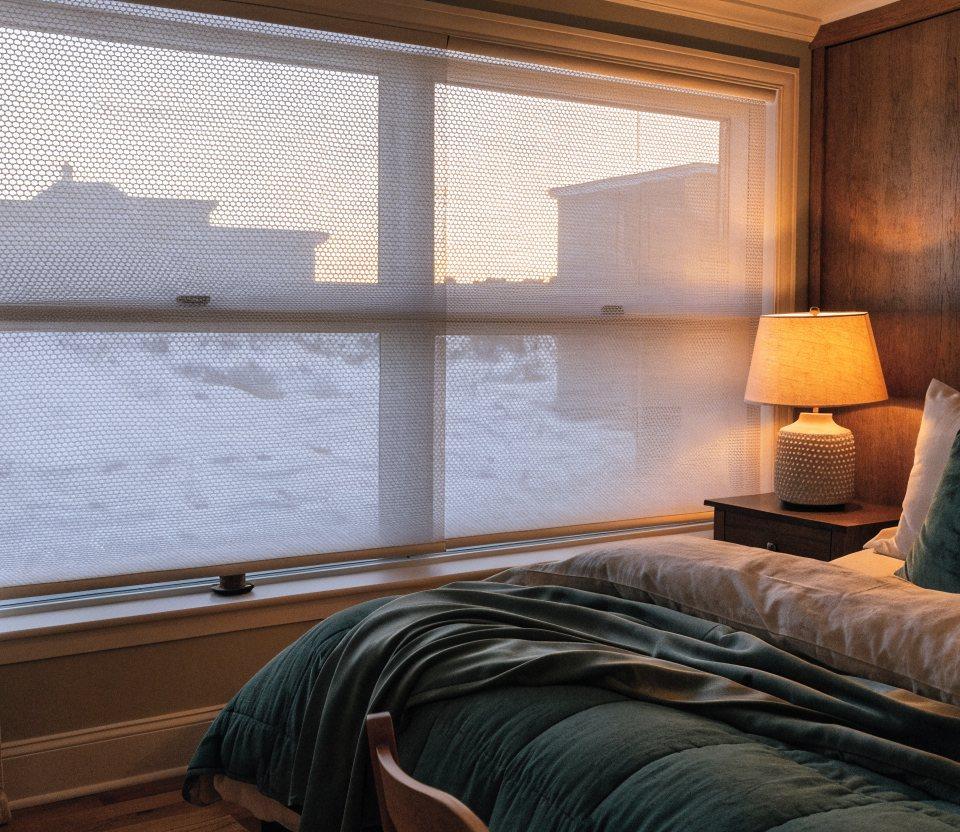 White cellular honeycomb shades half lowered over a bedroom window with snow outside and warm lamplight