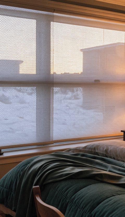 White cellular honeycomb shades half lowered over a bedroom window with snow outside and warm lamplight