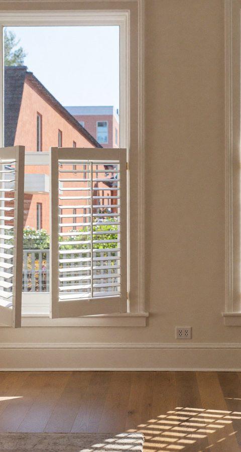White plantation shutters with louvers tilted open on tall windows in a sunlit Denver living room with oak floors
