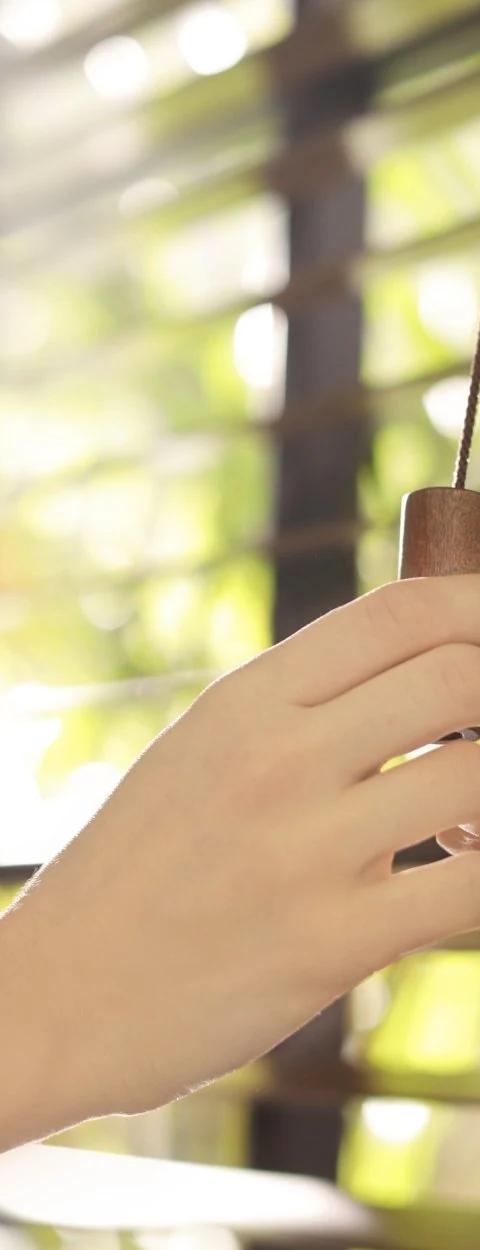 Hand adjusts dark horizontal blinds at a sunlit window