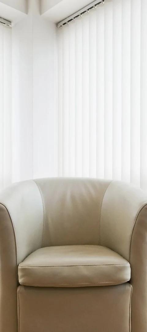 White vertical blinds cover a large window beside a pale armchair