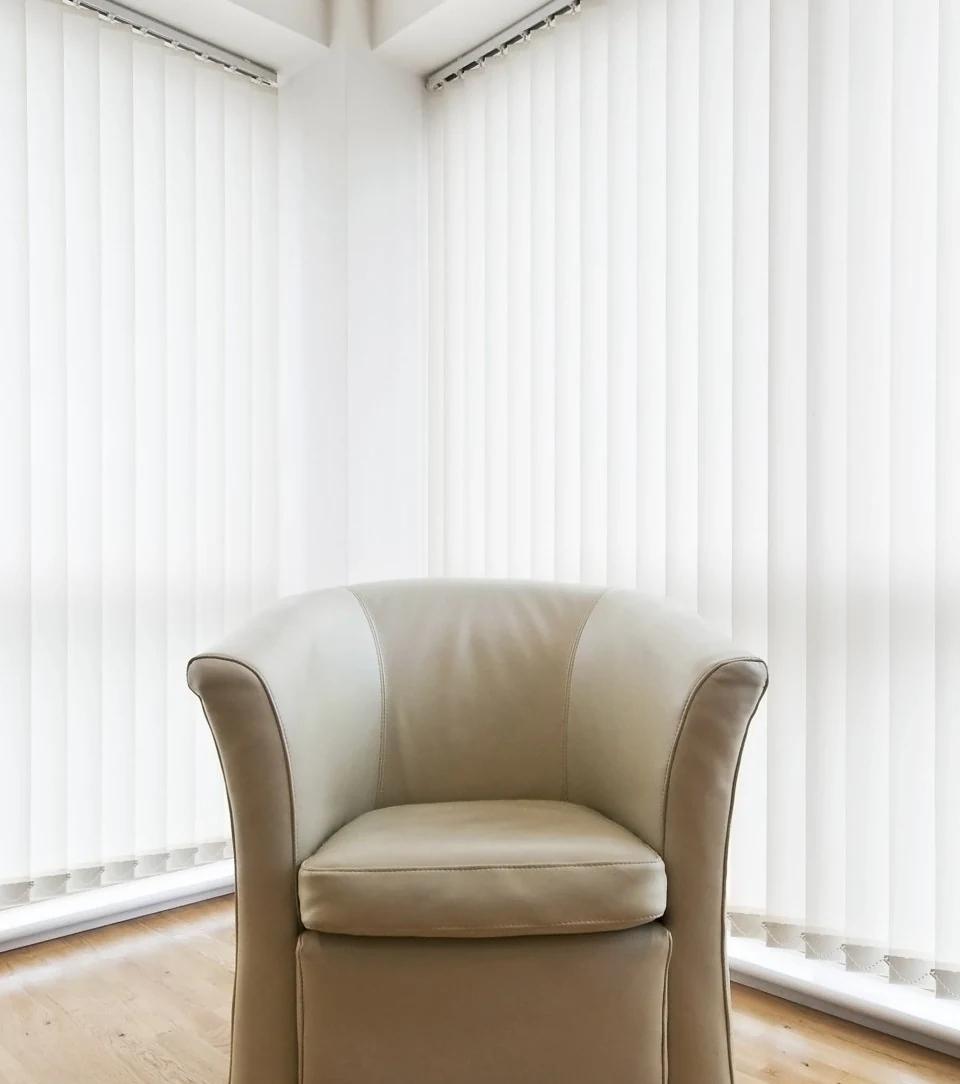 White vertical blinds cover a large window beside a pale armchair