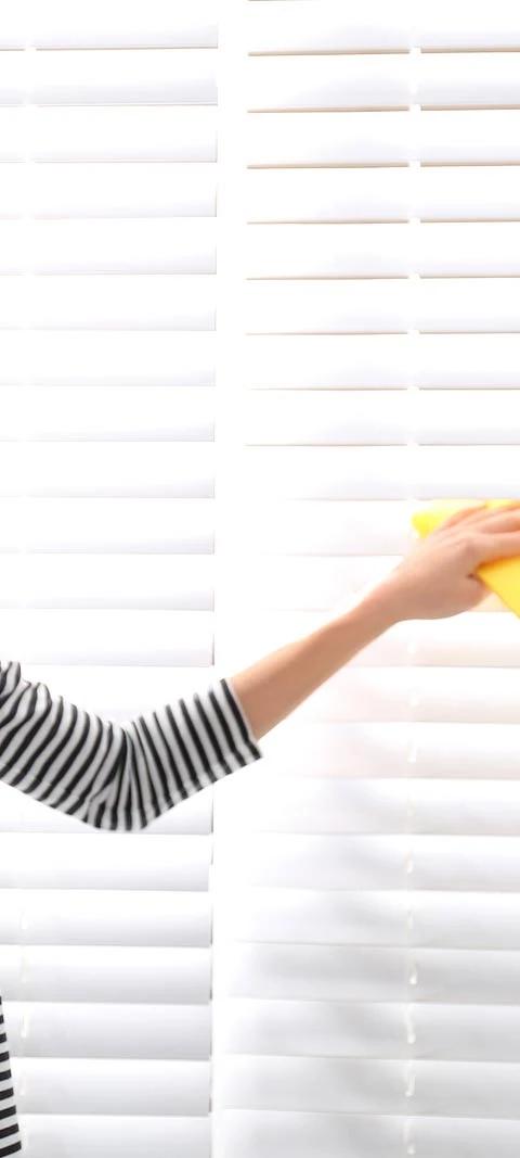 Woman cleans white horizontal blinds with a cloth beside a window