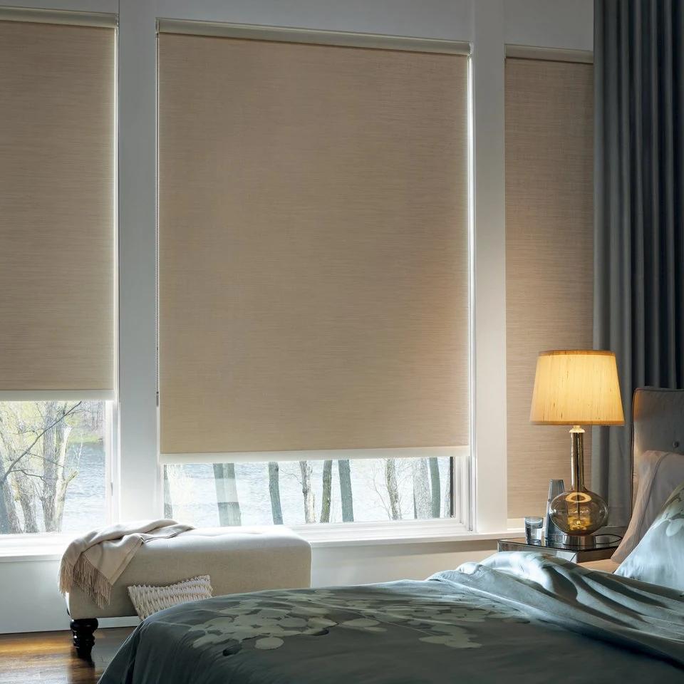 Roller shades partly cover two tall windows beside a bedroom lamp